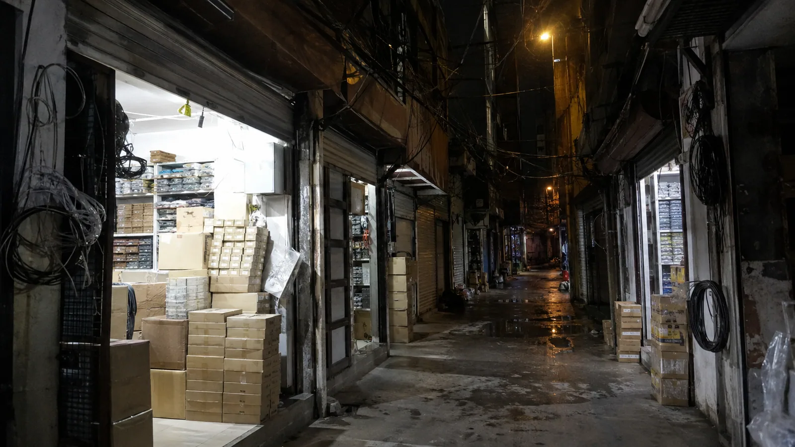 An electronics market alley at night — boxes, cables, half-open shutters