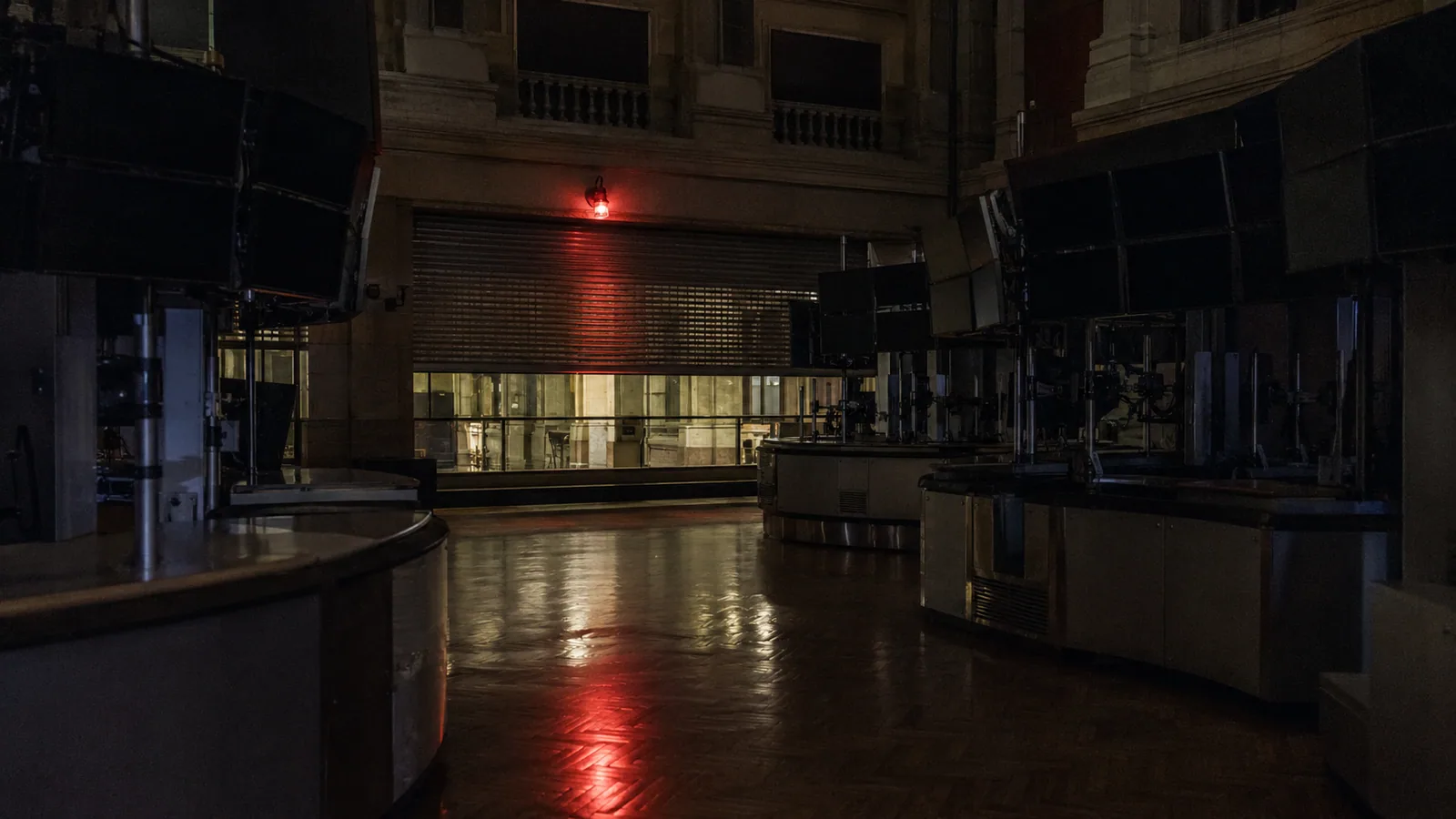 An exchange hall frozen mid-session, wall displays dark and a red beacon reflecting off the floor