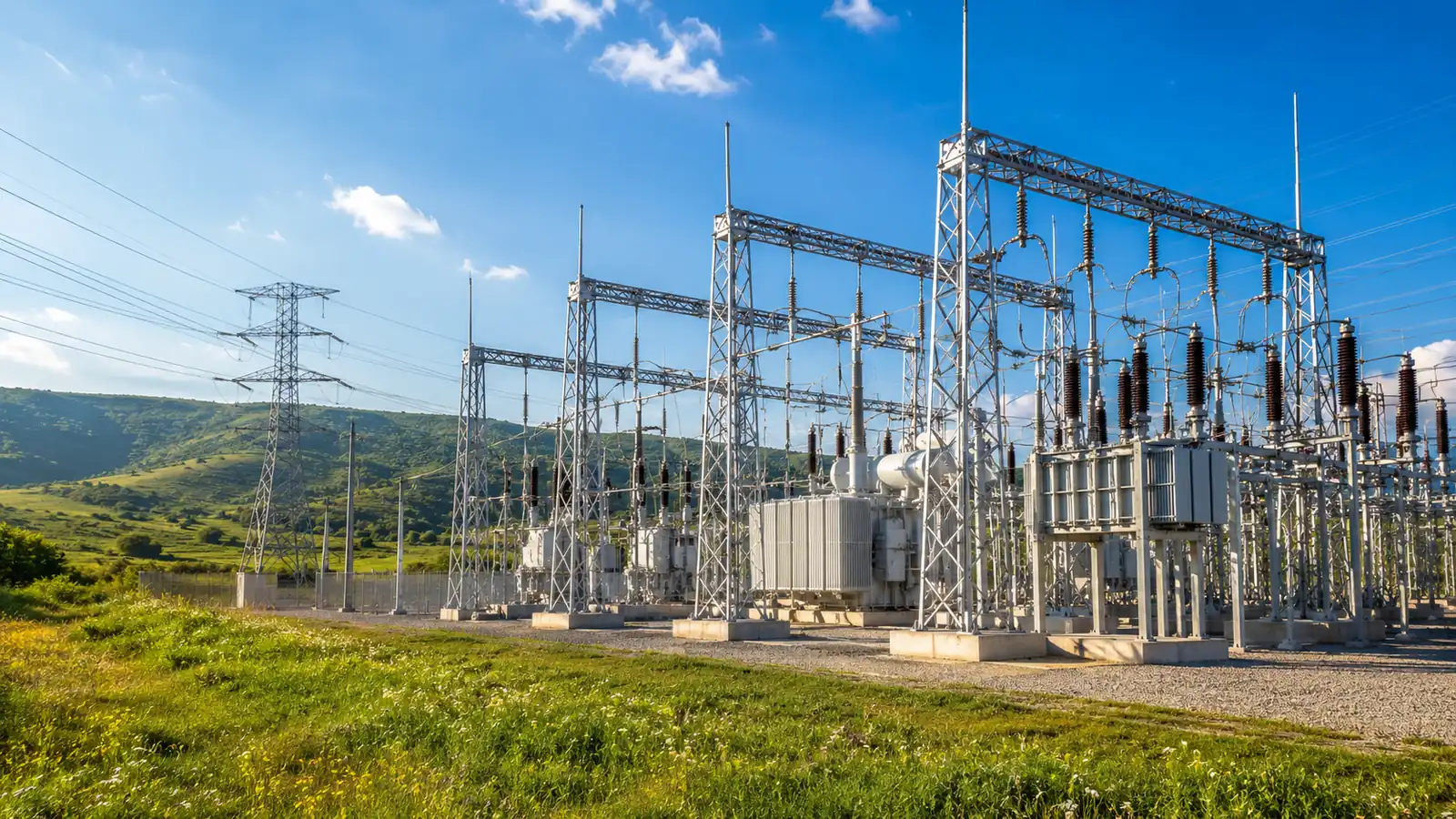 An electrical substation and transmission towers under a clear blue sky, with green grass below and hills in the distance
