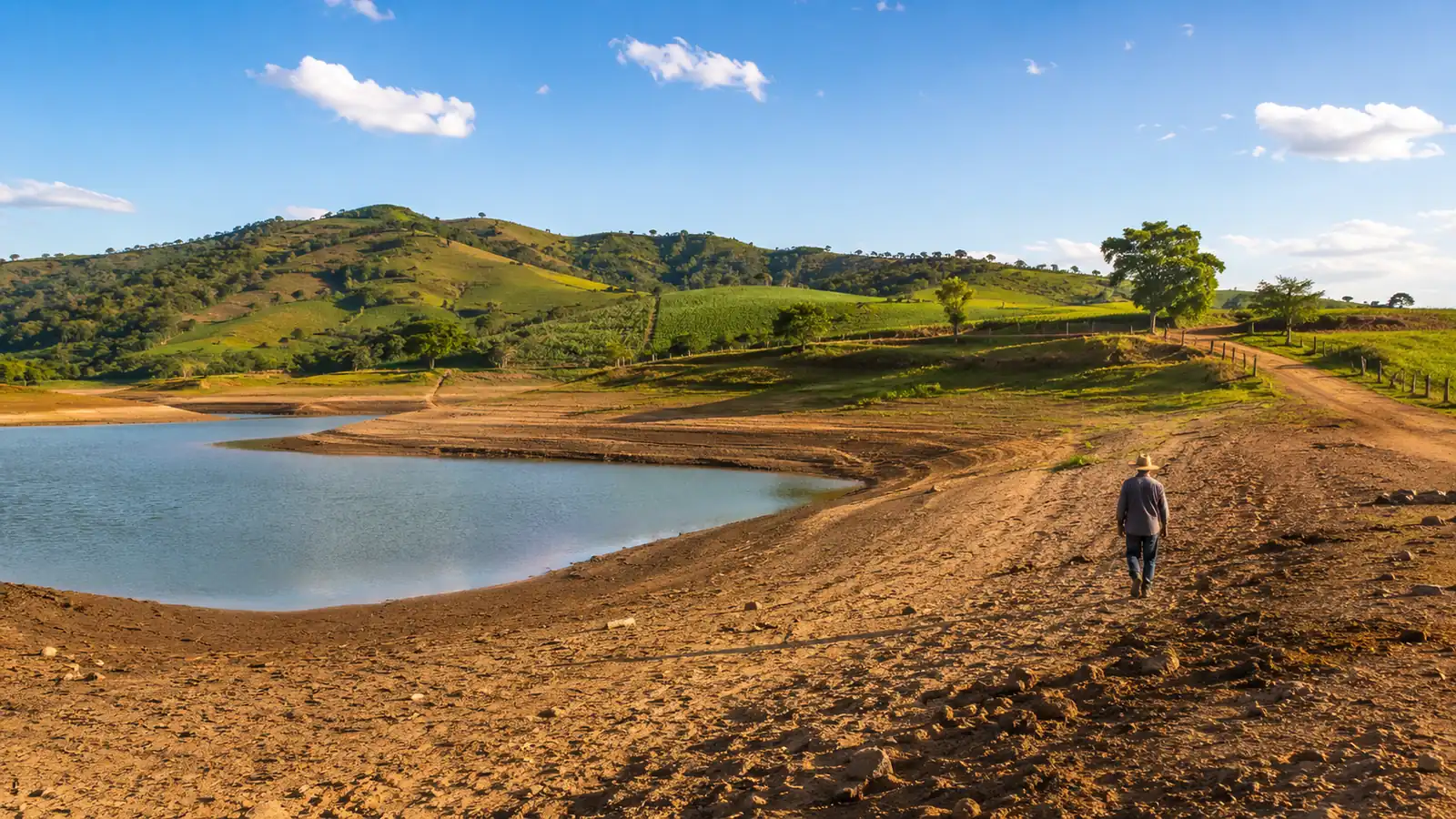 A rural reservoir with a low water line in afternoon light, exposed banks and green hills behind