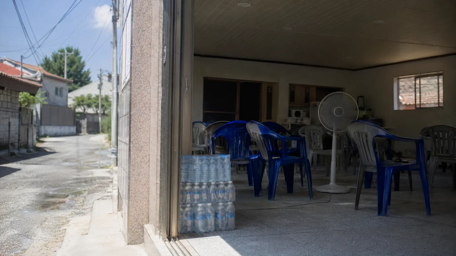 A cooling shelter doorway on a scorching afternoon, fan turning and bottled water stacked inside