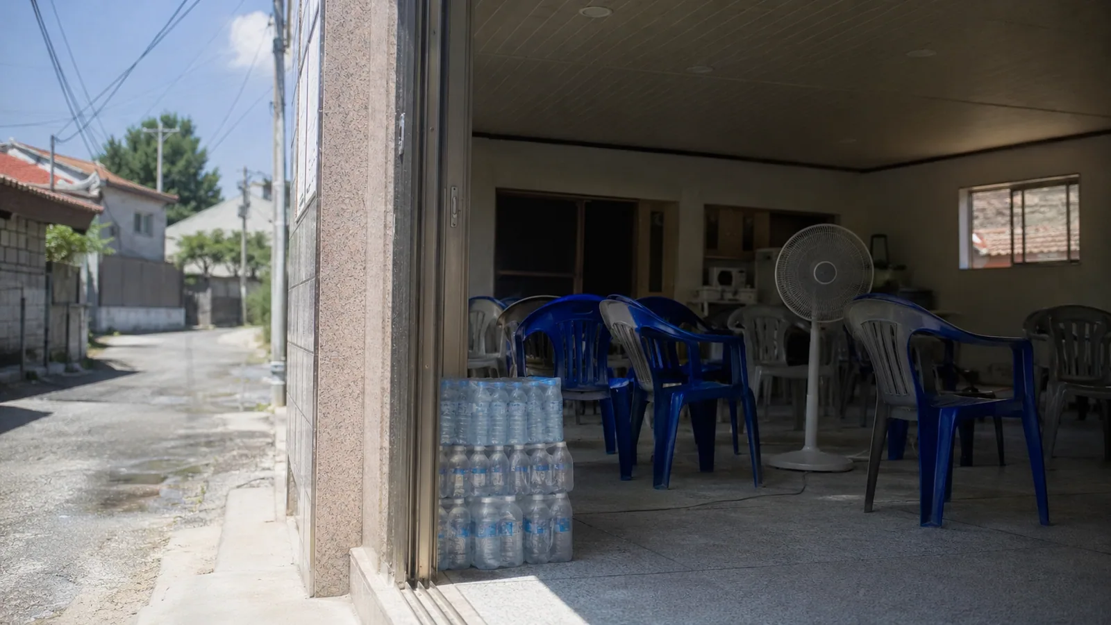 A cooling shelter doorway on a scorching afternoon, fan turning and bottled water stacked inside
