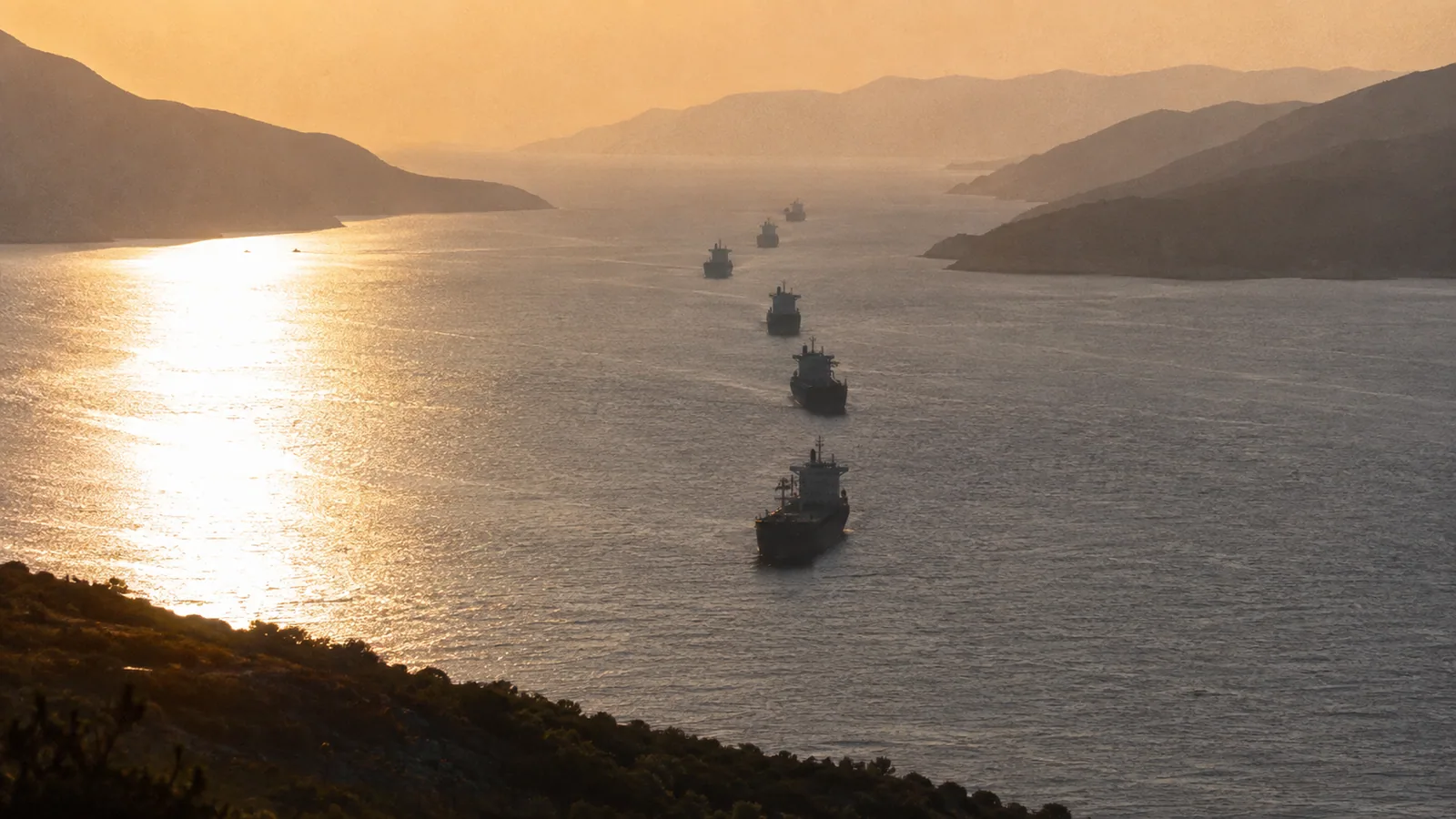 Tankers in single file threading a narrow strait between hazy shores at golden hour