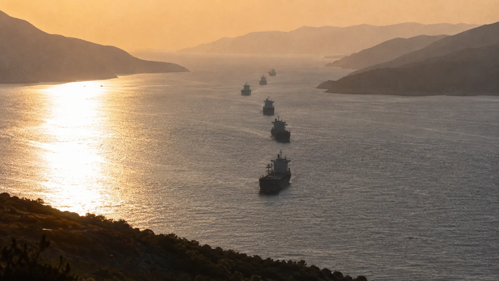 Tankers in single file threading a narrow strait between hazy shores at golden hour