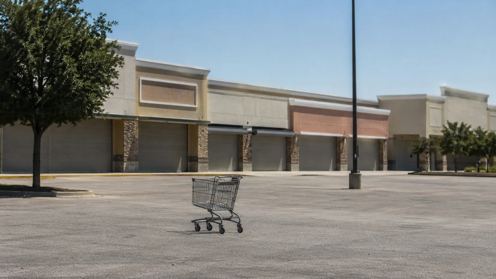 A strip mall at midday — shutters down, one cart alone on the asphalt