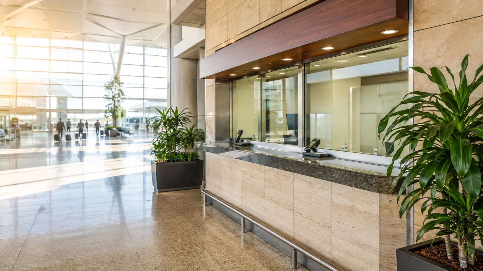 A currency exchange counter in a sunlit airport concourse with potted plants and travelers passing in the distance